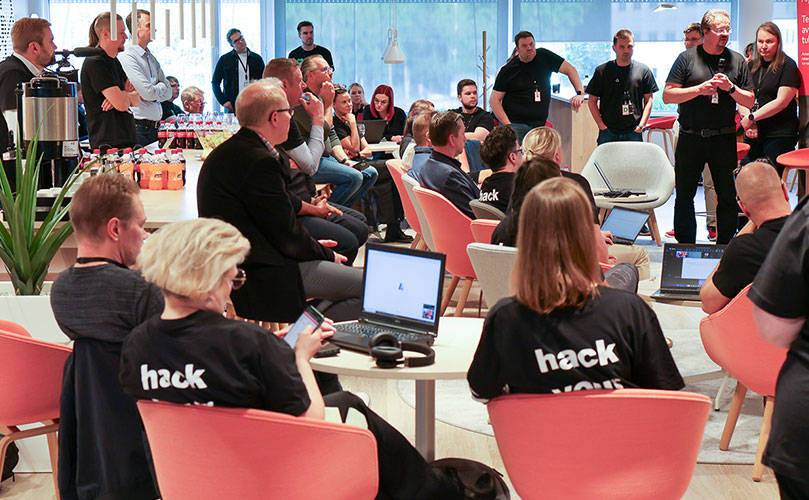 Suuri ihmisjoukko huoneessa istumassa, heillä on päällään t-paidat, joissa lukee 'hack'