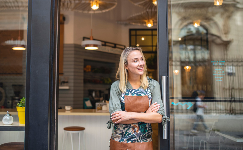 Una mujer en un delantal apoyada en una puerta de una cafetería mirando hacia un lado y sonriendo