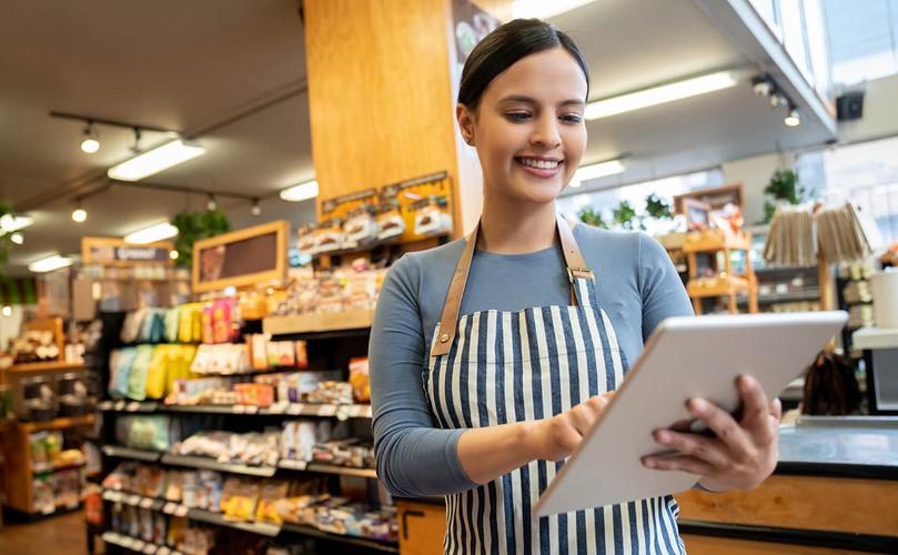 A happy supermarket employee looking to her tablet with aisles full of groceries behind her