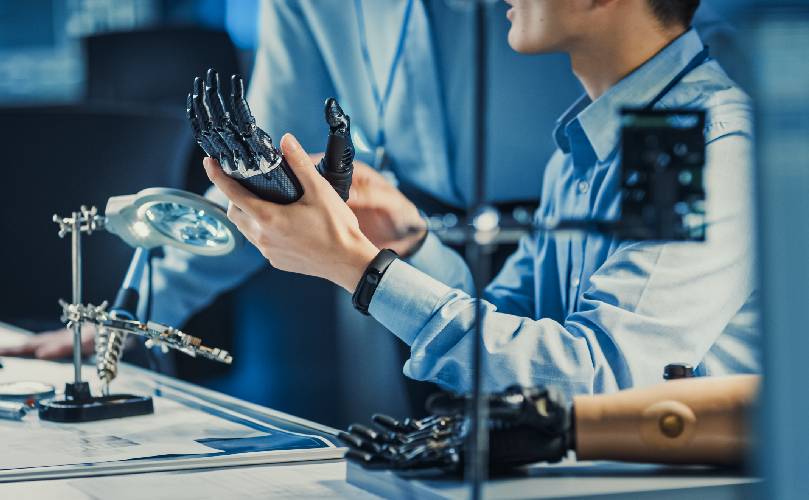  partial picture of a man showing a robotic left hand to another person, and the right robotic hand is on the table next to him