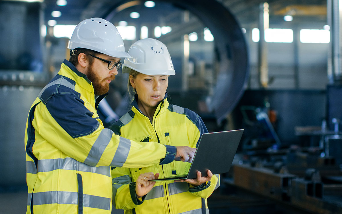 Male and female industrial engineers in hard hats discuss while using laptop Manufacturing plant