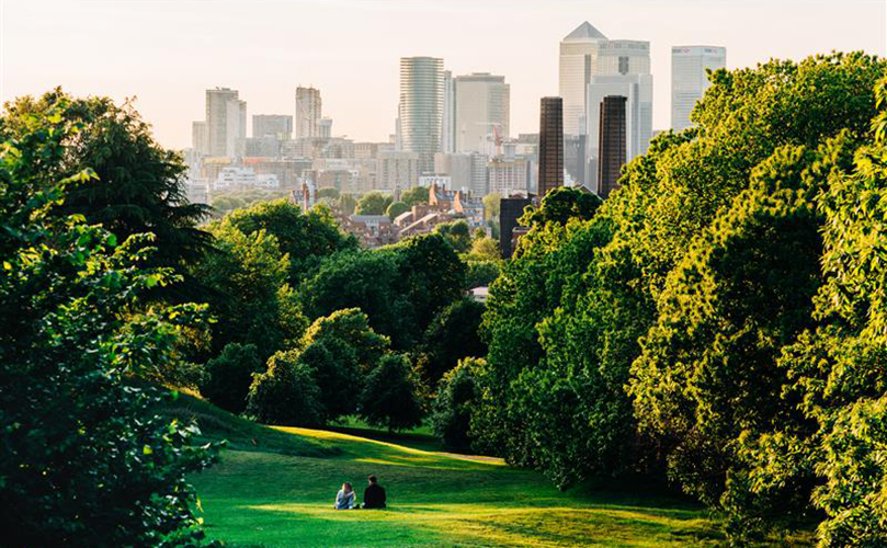 A couple enjoys a walk in a park, with a city skyline rising in the background.