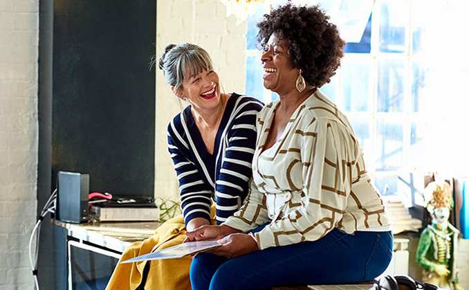 Two women sitting on a bench in a well-lit room, engaged in conversation and smiling at each other.