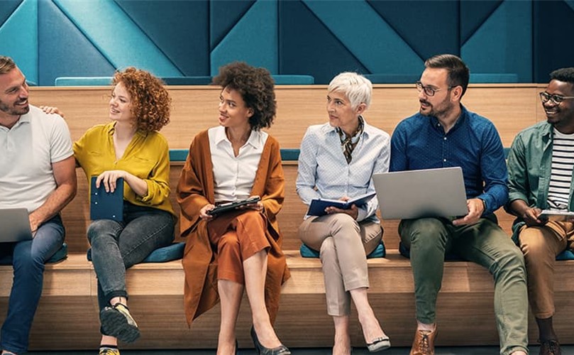 A diverse group of six individuals seated on wooden benches, engaged in a discussion, with laptops and notebooks in hand.