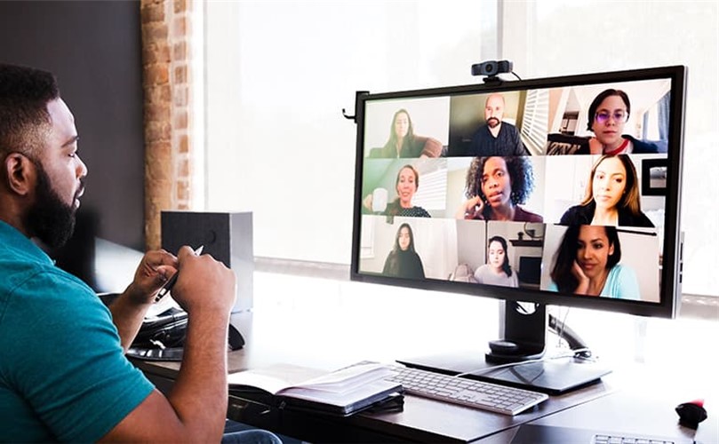 A man at a desk interacts with several people during a video call on his computer.