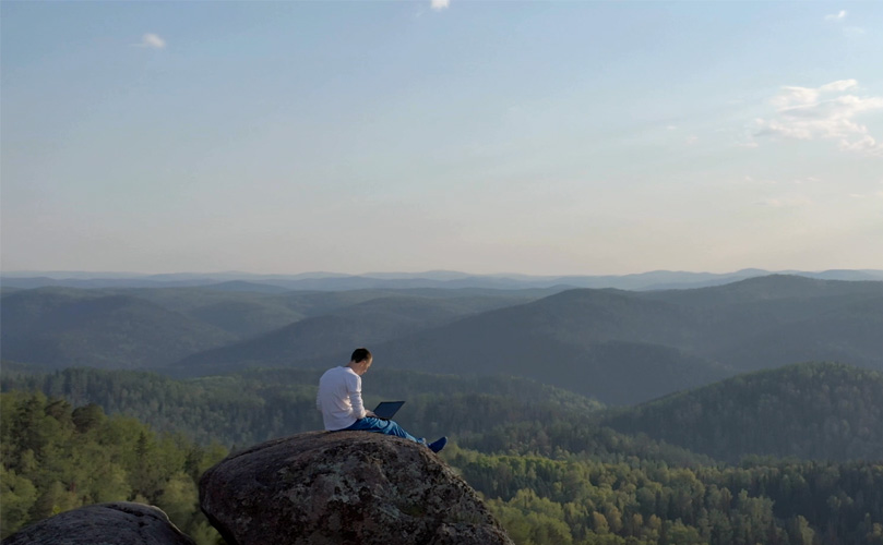 A man on a rock, using a laptop, surrounded by nature