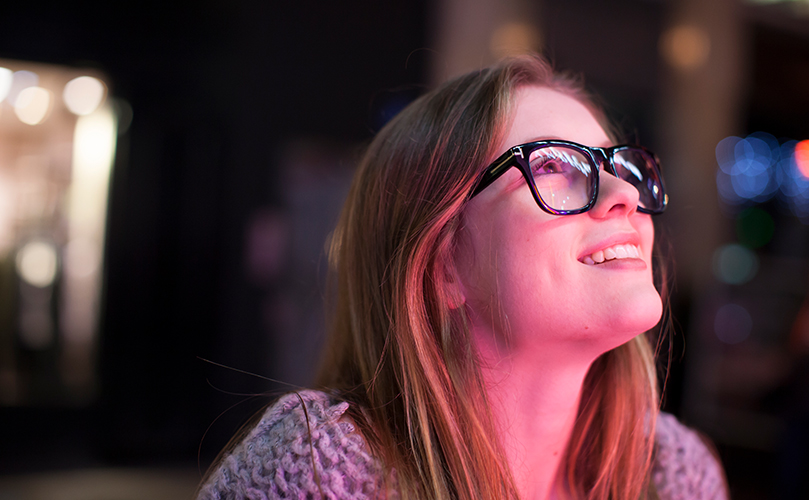 A woman with glasses gazes upward, observing the sky with a thoughtful expression.
