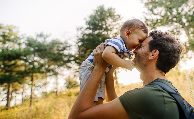 A man gently holds a baby in a lush green field, surrounded by nature under a clear blue sky.