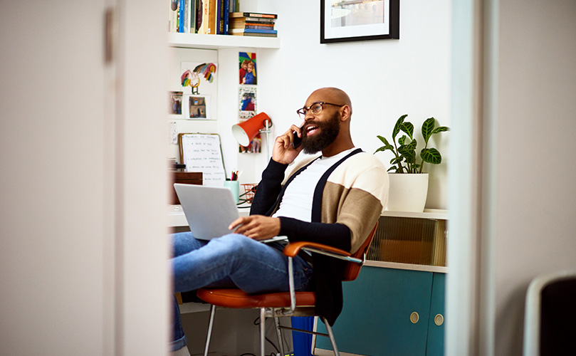 A man sitting with a laptop on his lap, conversing on the phone while seated in a chair.