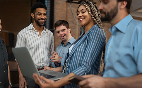 Two men and one woman dresssed in business wear are stood together. One man has Down Syndrome. The woman holds a laptop; they are all looking at it and smiling.