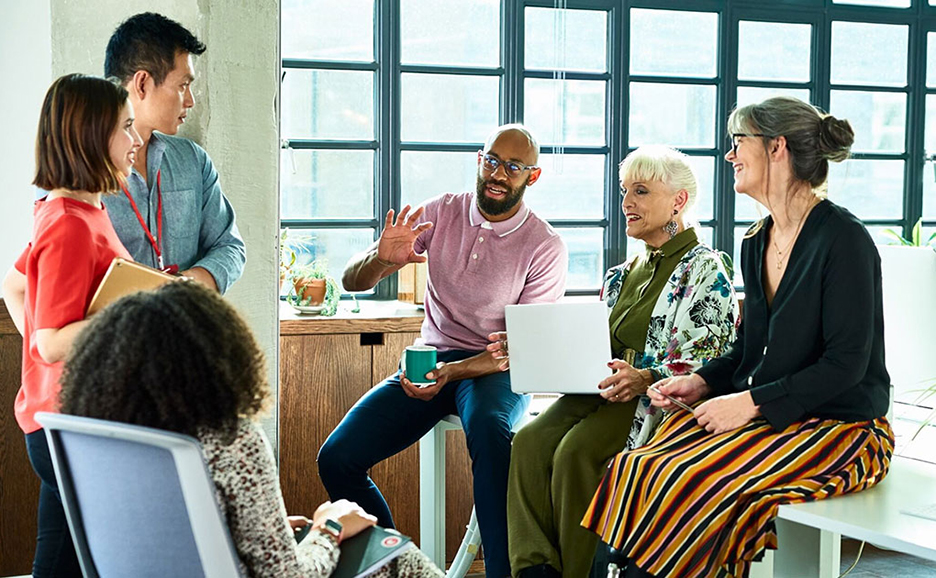 Six diverse people are working casually together at their desks in an office. One person holds a laptop that they are all looking at.