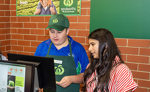 A young person in a green Woolworths uniform practicing on a Fujitsu register. They are guided by a mentor.