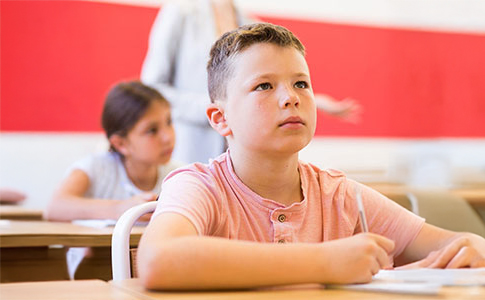 A white male child is sat in a classroom looking up with an expression of concentration. Sat behind him is a female child at a desk and an adult standing up.