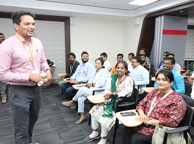 Visually impaired female and male Indian students sat in rows at lecture tables. A male lecturer stands at the front of the class.