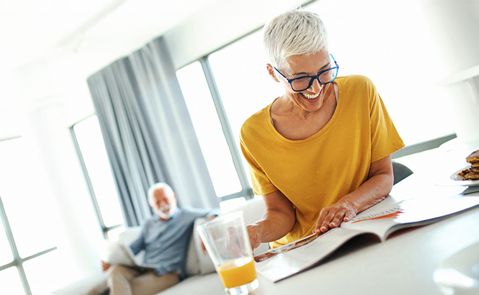 笑顔の女性が雑誌を読み、手前にオレンジジュースとパンケーキ。背景に男性がソファに座っている。