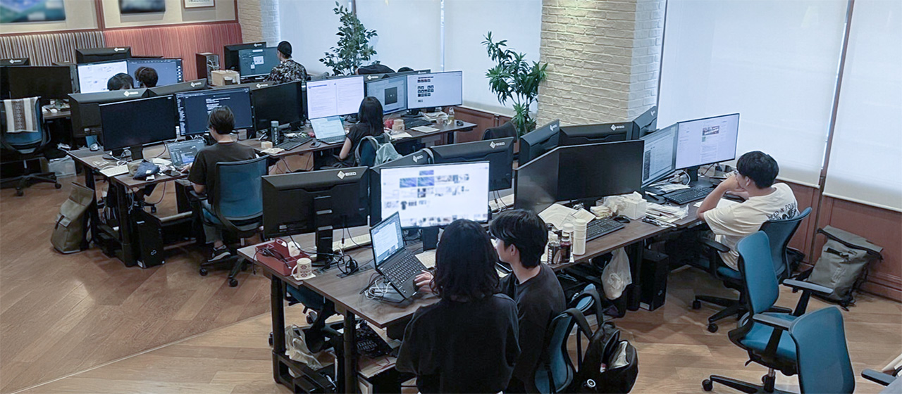 オフィス内で複数のスタッフがデスクに並び、複数のモニターを使ってパソコン作業をしている様子。開放的な空間に観葉植物やレンガ調の柱が配置され、チームで集中して働く雰囲気が伝わる。