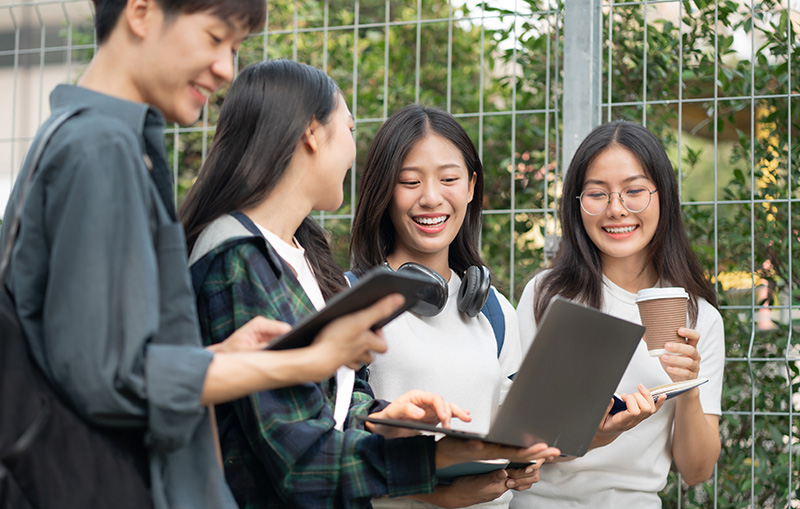 屋外でノートパソコンやタブレットを見ながら笑い合う学生たち