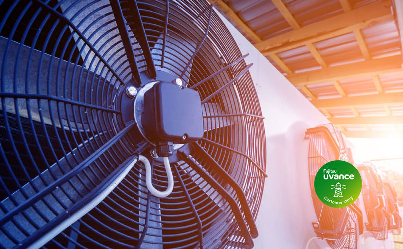 A wall-mounted air conditioner with a large fan, metal grille, and energy efficiency sticker against a blurred building roof and blue sky.