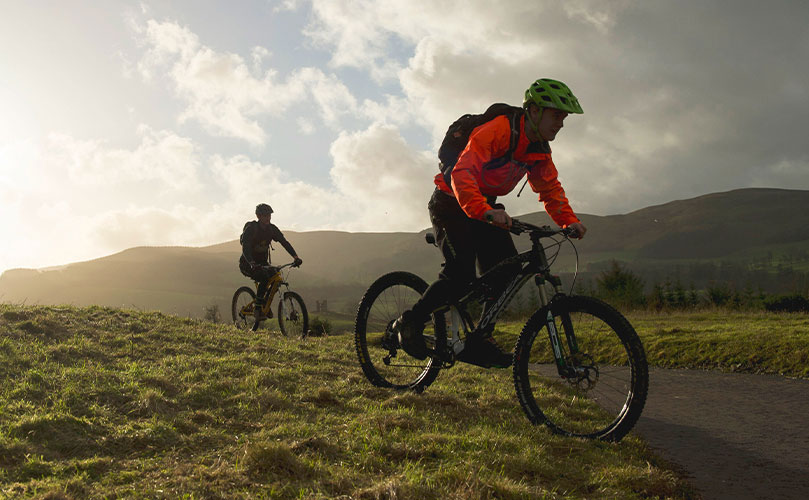 Two mountain bikers riding on a grassy trail with hills in the background under a partly cloudy sky.