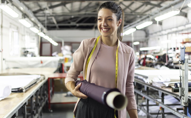 A woman walking and smiling with a measuring tape around her neck and holding a roll of fabric