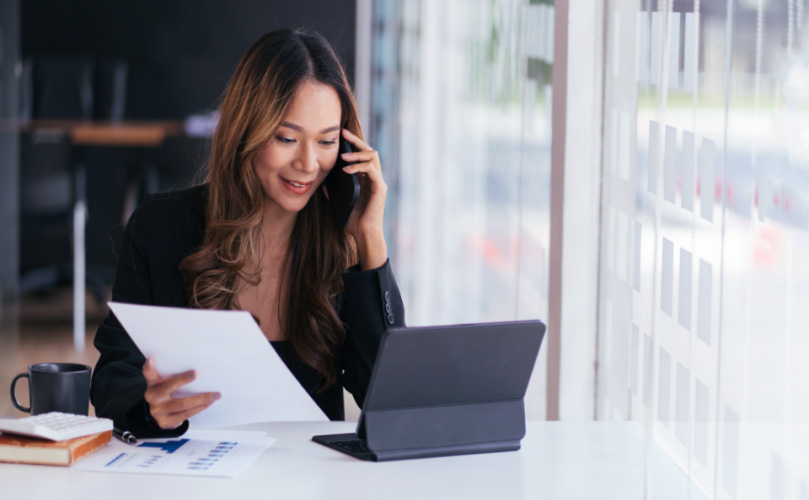 A woman sitting at her desk chatting on the phone while holding a piece of paper and looking at a tablet