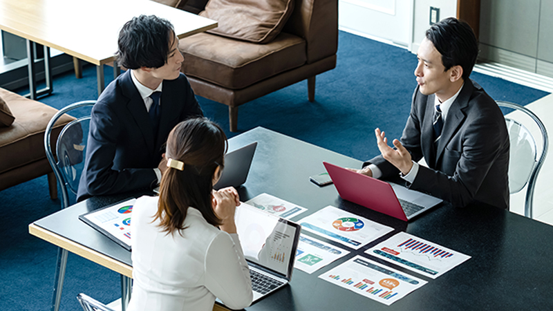 Three people with computers in front of them sitting at a desk and chatting