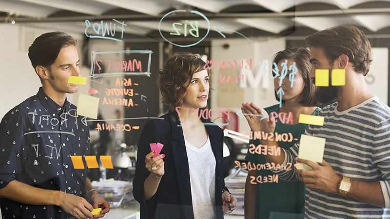 Four professionals discussing and writing on a glass wall covered in sticky notes and marker notes in an office setting