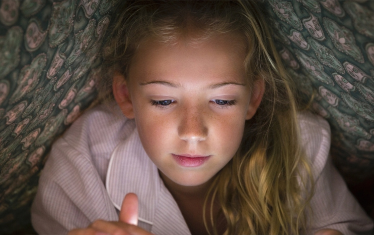 A young girl lies on a bed, her hand resting on her phone as she engages with the device.