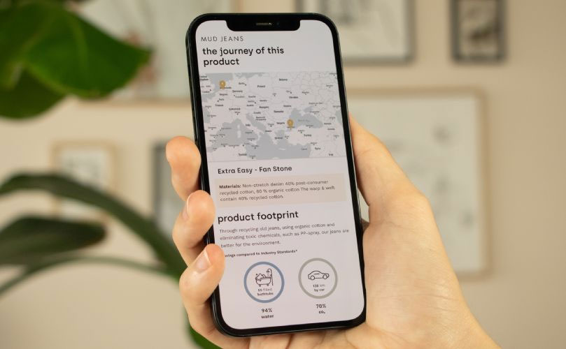 A hand holds a smartphone displaying a webpage titled the journey of this product with a map, product info, and icons showing the environmental footprint