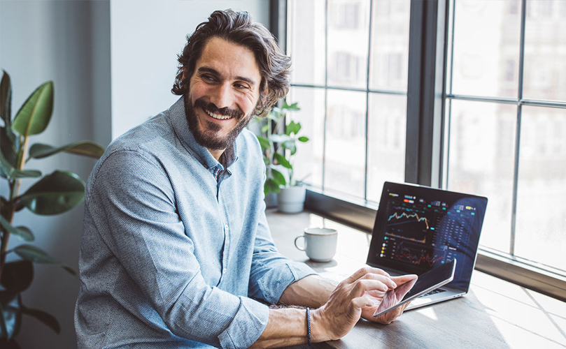 A man sitting a desk with a computer is looking back while smiling and holding a tablet.