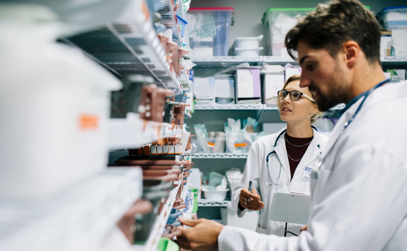 A man and a woman in lab gear are looking at a shelf of medical supplies