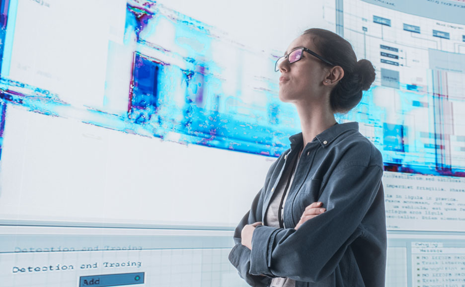 A woman stands with folded arms, contemplating data on a large interactive digital display.