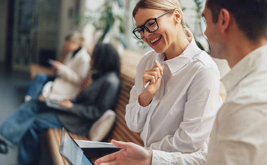 Female employee with a device, smiling and conversing with a male colleague in an office.