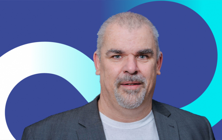 A bearded man in a suit jacket stands confidently against a blue and white background.