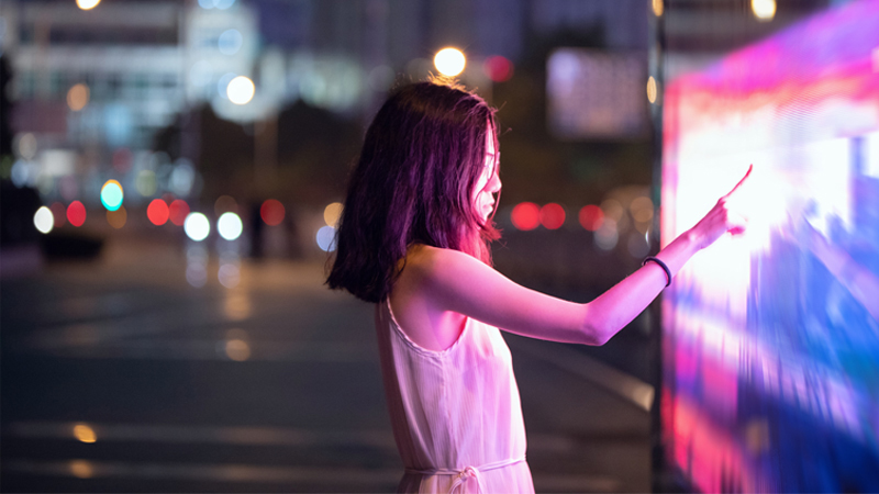 A woman in the middle of the street touching a neon digital screen