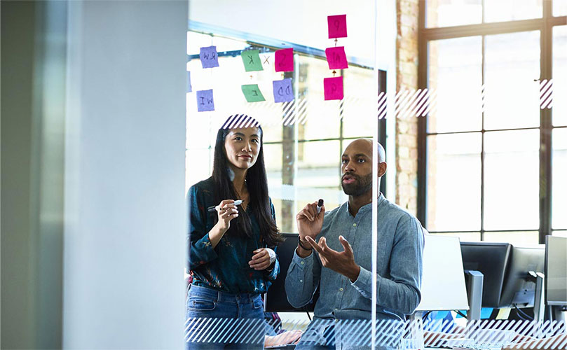 Male and female colleagues discussing ideas with sticky notes on a glass board.