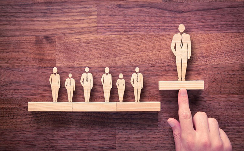 Auf einem Holztisch stehen 6 kleine und eine große Holzfigur von Menschen. Eine Hand zeigt auf die große Holzfigur