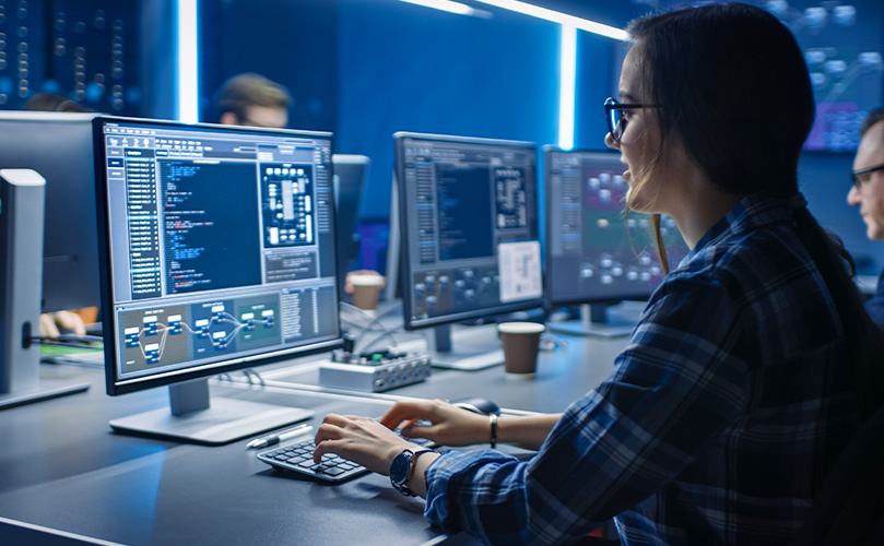 Specialist analyzing cybersecurity data and system performance on multiple computer monitors in a control room