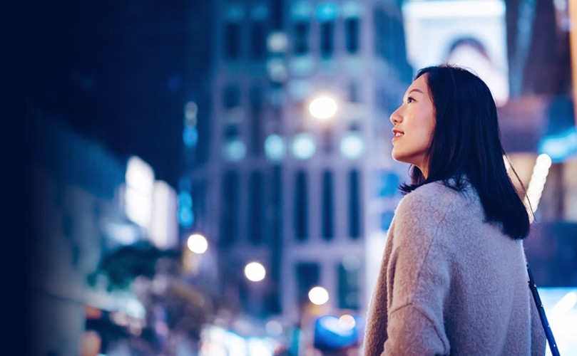 A woman stands in a city street at night, framed by illuminated buildings and a dark sky.