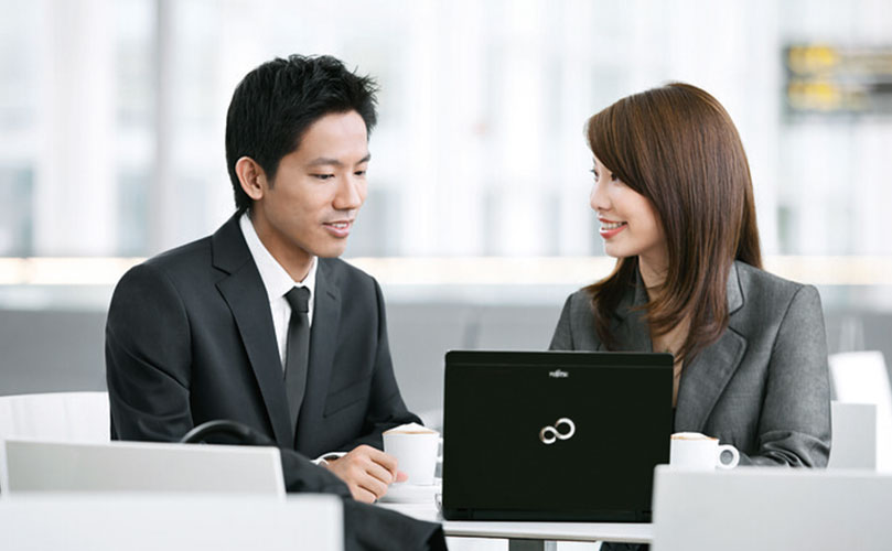 Zwei Kollegen sitzen an einem Tisch mit einem Laptop und Kaffeetassen und scheinen etwas in einem hellen Büro zu besprechen.