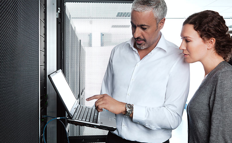 Zwei Kollegen in einem Serverraum prüfen Informationen auf einem Laptop, während daneben Serverracks und Netzwerkkabel sichtbar sind.