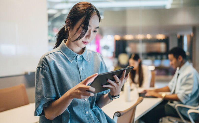 Eine junge Frau arbeitet im Büro an einem digitalen Tablet. Im Hintergrund arbeiten unscharf Kollegen weiter.