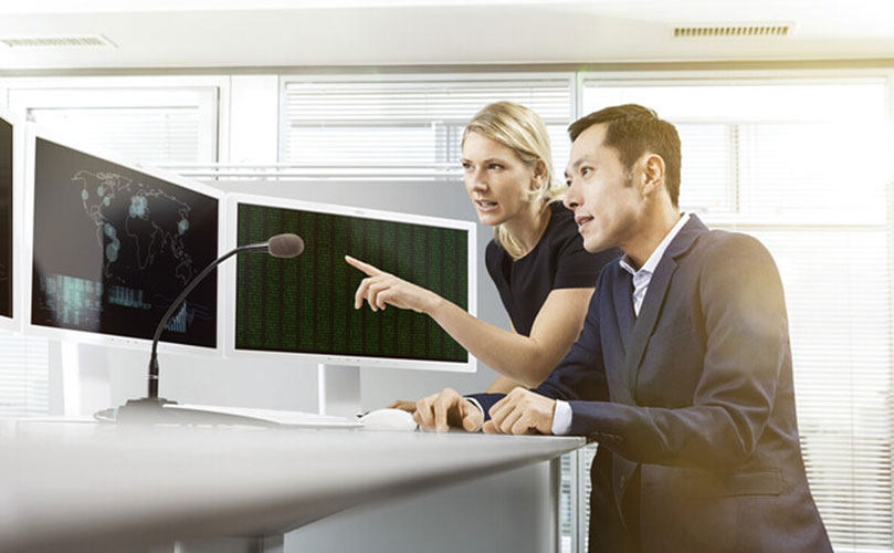 Zwei Kollegen arbeiten an einem Schreibtisch mit mehreren großen Monitoren, wobei ein Kollege auf einen Bildschirm mit Daten zeigt.