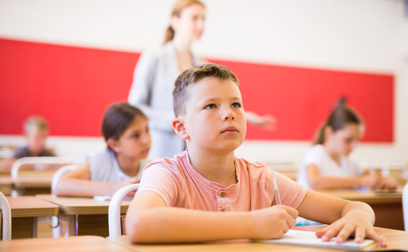 Children sitting at desks in a classroom, with a teacher standing in the background.