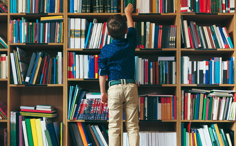 A child in front of book shelf reaching for a book at the top