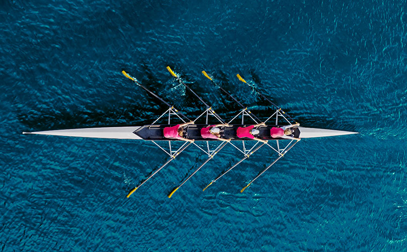 Aerial view of a four-person rowing team in a boat, rowing on clear blue water.