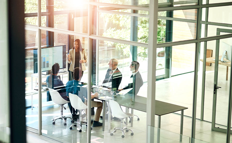 ガラス張りの会議室で、女性がプレゼンテーションをしている。