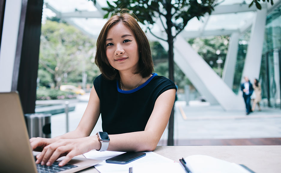 ノートパソコンで作業するアジア人女性。屋外のカフェで笑顔