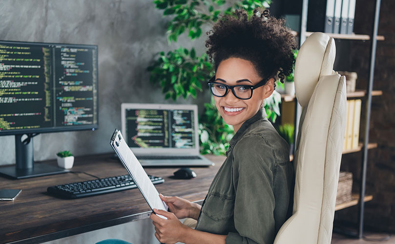 A woman smiling at the camera while sitting at a desk with computers