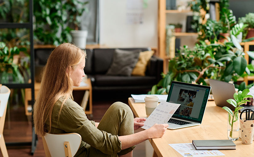 A woman is seated on a wooden floor, holding a document in one hand while looking at a laptop screen.